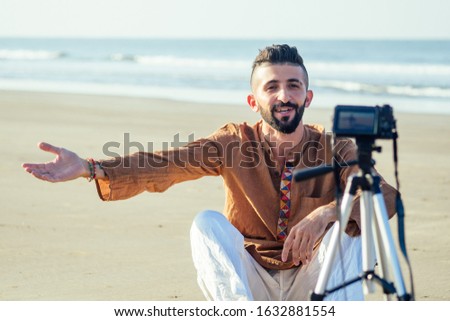 arabian man sitting on sand and talking tripod video making in beach. Travel videographer lifestyle content maker concept