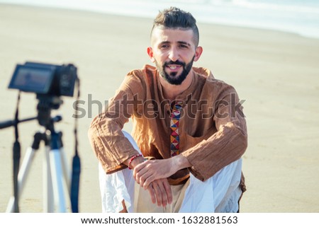arabian man sitting on sand and talking tripod video making in beach. Travel videographer lifestyle content maker concept