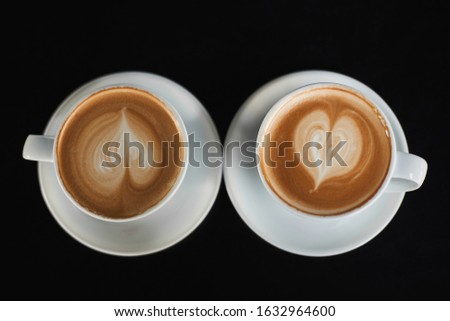 Two white cups of Cappuccino coffee with heart-shaped milk foam. Top view on two cups of latte coffee with heart figure on milk foam isolated on black background. Concept of breakfast. View from above