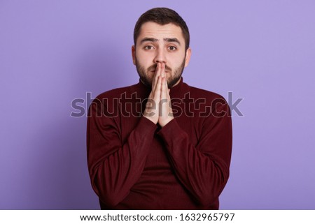 Close up portrait of bearded handsome man with hopeful facial expression putting hands together, wearing dark red sweatshirt, being nervous, looking directly at camera. People and emotions concept.