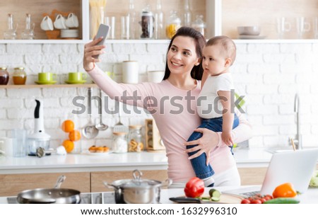 Cheerful mom with baby taking selfie on cellphone at kitchen, empty space