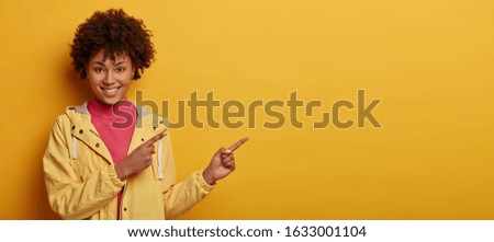 Check this out. Photo of attractive smiling woman with Afro hair points on copy space, has satisfied face expression, wears yellow anorak, isolated over yellow background, smiles with teeth.