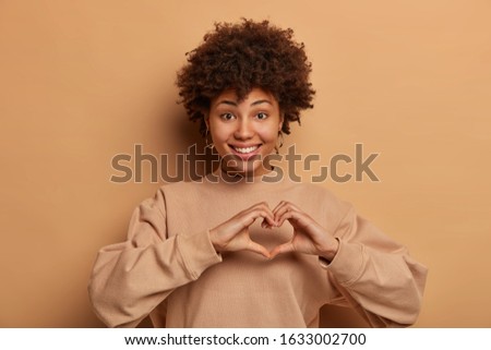 Smiling girlfriend shapes heart with both hands, expresses effection, care and love to husband, sends valentine, wear casual sweater, stands against brown wall. Lovely female makes love sign
