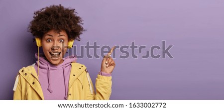 Millennial hipster girl with Afro hair enjoys music podcast in headphones, points fore finger on copy space for your advertising, has excited surprised happy expression, isolated on purple background