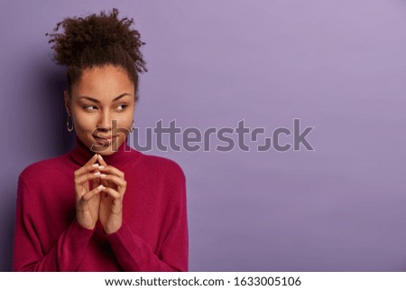 Mysterious curly haired woman keeps hands together with intention to do something, looks with intrigued expression aside, wears casual jumper, has interesting plan, isolated on purple background