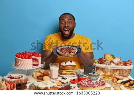 Positive bearded plump man holds plate with homemade pie, enjoys eating unhealthy but delicious desserts, sits at table overloaded by sweet products, doesnt keep to diet, wears yellow t shirt Royalty-Free Stock Photo #1633005220