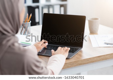 Unrecognizable muslim woman working on laptop with black screen, typing on keyboard, sitting in modern office, over shoulder view, mockup