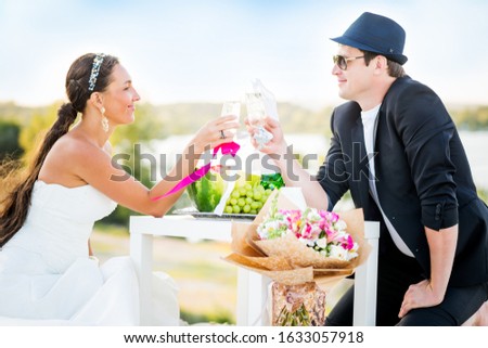 Side view of charming young couple bride and groom clink glasses of champagne while sitting at table with fruits and bouquet of flowers, background of river and the forest on a sunny warm summer day