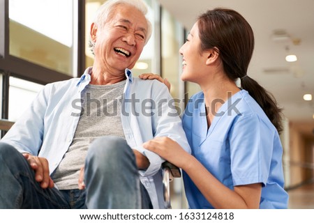 young friendly asian female caregiver talking chatting to happy senior man in hallway of nursing home Royalty-Free Stock Photo #1633124818