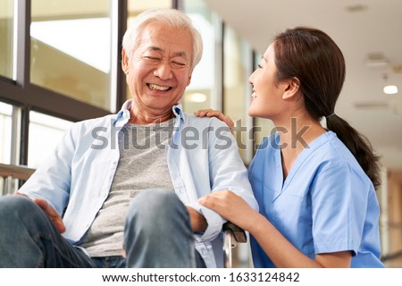 young friendly asian female caregiver talking chatting to happy senior man in hallway of nursing home Royalty-Free Stock Photo #1633124842