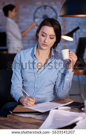 Young businesswoman sitting at desk, writing, drinking coffee.