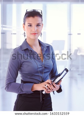 Young attractive brunette caucasian secretary standing at business office with personal organizer, looking at camera, smiling. Woman suit, copyspace.