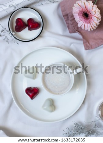 porcelain cup of coffee with cream and delicious bonbons with a heart shape on a white bed. good morning concept. flat lay, top view, vertical image