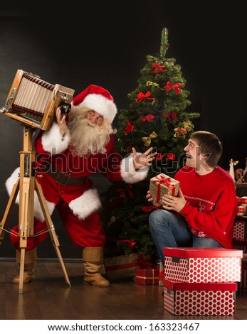 Santa Claus taking picture of cheerful man with old wooden camera at home near Christmas tree