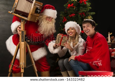 Santa Claus taking picture of couple with old wooden camera at home near Christmas tree