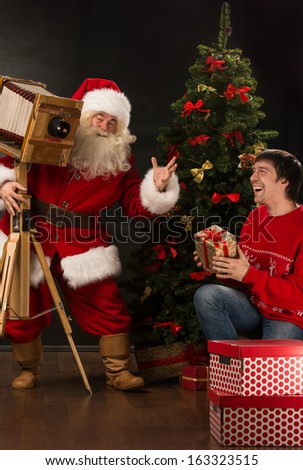 Santa Claus taking picture of cheerful man with old wooden camera at home near Christmas tree