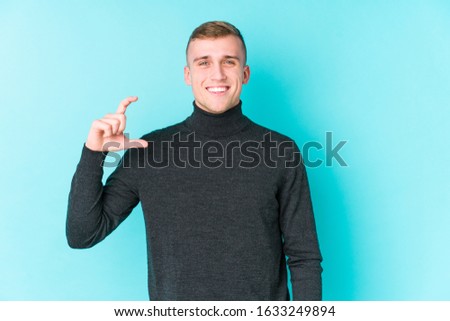 Young caucasian man on a blue background holding something little with forefingers, smiling and confident.