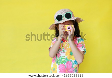 Asian little girl taking photo by toy camera. Child tourists in floral pattern summer dress and hat with sunglasses isolated on yellow background.