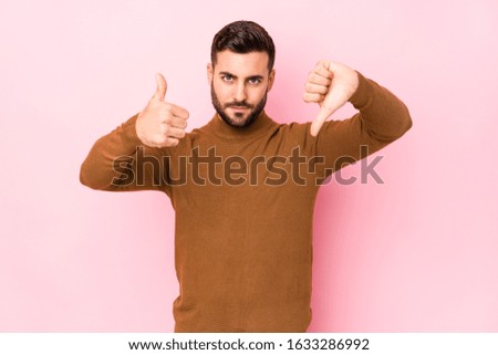 Young caucasian man against a pink background isolated showing thumbs up and thumbs down, difficult choose concept