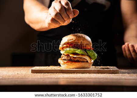 home made hamburger made by white sesame bun, tomato slice, salad, cheese, grilled meat and onion on wooden tray wooden table with cooking decorate chef hand in dark isolate background stock photo