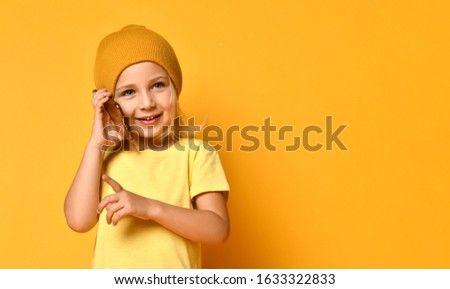 Little smiling blond girl in yellow t-shirt and hat talking by phone and looking aside over yellow background. Trendy children outfit concept