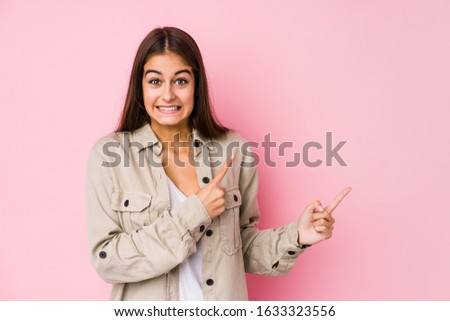 Young caucasian woman posing in a pink background shocked pointing with index fingers to a copy space.
