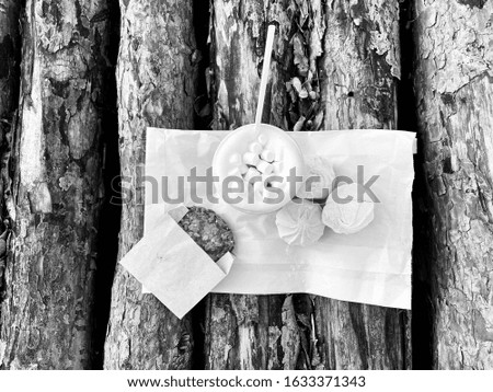 A paper cup of latte with marshmallow and an oat cookie on wooden logs black&white