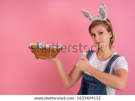 Easter holiday concept. Pretty young woman in bunny ears showing thumbs ups and holding a basket with Easter colored eggs isolated on pink background