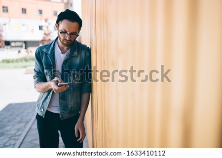 Trendy dressed blogger 20s checking received smartphone notification concentrated on update, Caucasian millennial in classic eyeglasses connecting to internet for browsing website on cellphone
