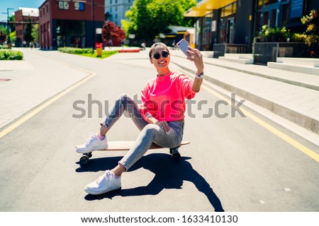Asian female influencer shooting video vlog for communicate with followers about skate sport sitting on longboard and smiling at front smartphone camera, millennial blogger taking selfie pictures