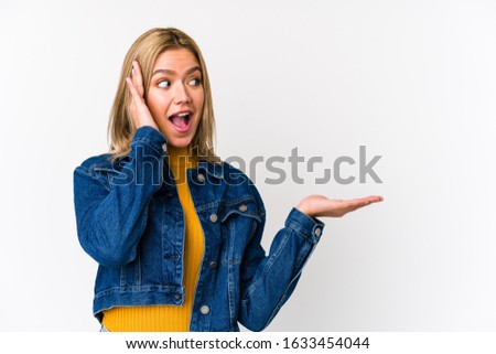 Young blonde caucasian woman isolated holds copy space on a palm, keep hand over cheek. Amazed and delighted.