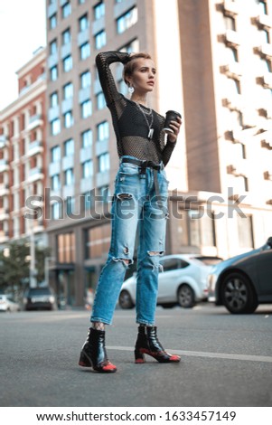 Young stylish woman standing outdoors on road urban background holding cup of hot coffee looking forward touching hair thoughtful
