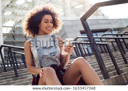 Young stylish woman in the city street sitting on concrete stairs holding smartphone looking up smiling dreamful