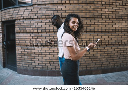Side view of happy millennial African American female holding smartphone and looking at camera while standing against brick wall with and man checking message on mobile phone in background