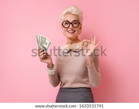Smiling grandmother shows money cash and makes okay gesture, demonstrates that everything is fine. Photo of kind elderly woman wears eyeglasses on pink background.