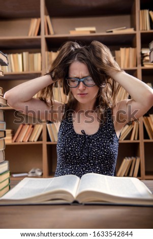 Young girl student reads a textbook sitting at a table with many books in the library