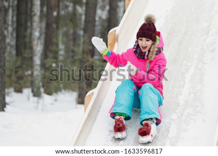 Woman slide down an icy wooden hill on a snowy day. Pretty girl emotionally laughs and rejoices in the winter outdoors. Bright clothes, high mountain, pine forest.