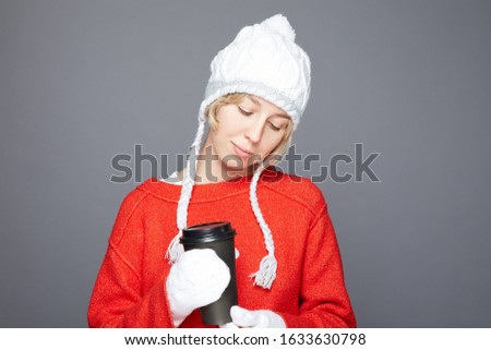 Portrait of happy Caucasian woman smiles pleasantly, keeps eyes closed, holds black cup of coffee, enjoys aromatic drink, wears white hat and red knitted sweater, isolated over grey background.