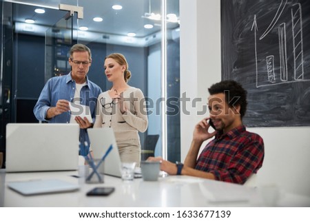 Gentleman showing notes to young woman while their coworker talking on cellphone stock photo
