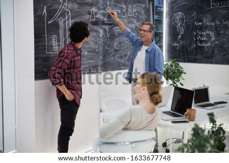 Cheerful man writing on chalkboard and chatting with colleagues stock photo