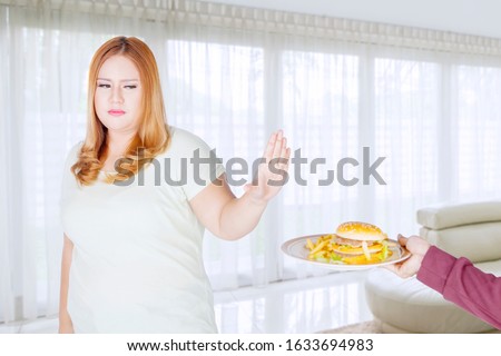 Portrait of obese woman rejecting to eat burger or unhealthy food at home. Dieting concept