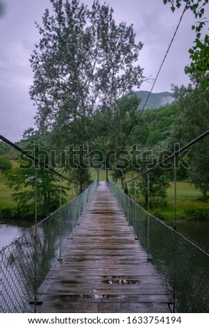 A vertical picture of a wooden bridge on a river surrounded by greenery with hills on the background
