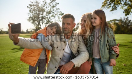 Portrait of cheerful parents with children taking pictures using smartphone in the park on a sunny day. Family, parenthood, and leisure concept. Horizontal shot. Front view