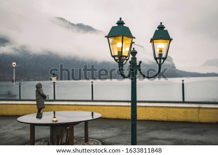 Foggy morning waterfront walking area of Austria highland Alps mountains lake and street lantern yellow illumination with unfocused person silhouette background 