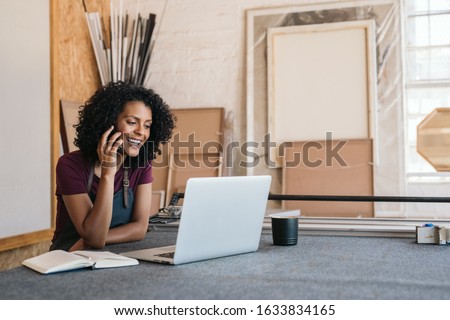 Young female artisan working on a laptop and talking with a client on the phone in her workshop