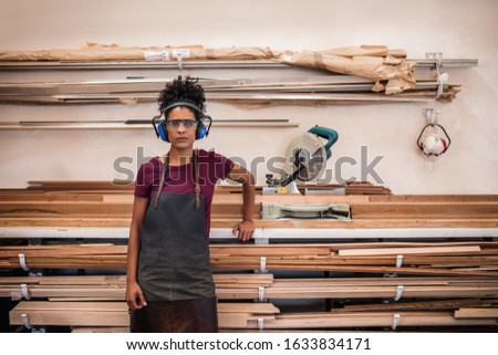 Portrait of a young female artisan in safety gear standing by wood and a saw in her picture framing workshop