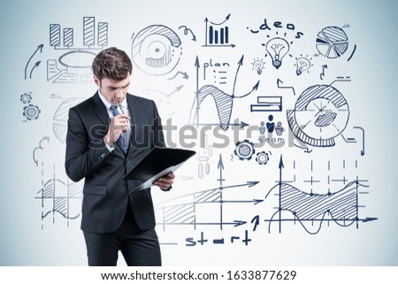 Pensive young businessman looking at clipboard standing near gray wall with business plan sketch drawn on it. Concept of brainstorming