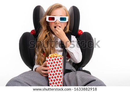 A little girl with makeup and long blonde hair sits on a car baby chair and looks film or cartoon with popcorn isolated on white background