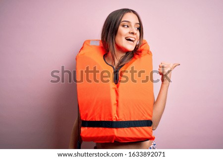 Young beautiful brunette woman wearing orange lifejacket over isolated pink background smiling with happy face looking and pointing to the side with thumb up.