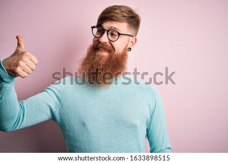Handsome Irish redhead man with beard wearing glasses over pink isolated background Looking proud, smiling doing thumbs up gesture to the side